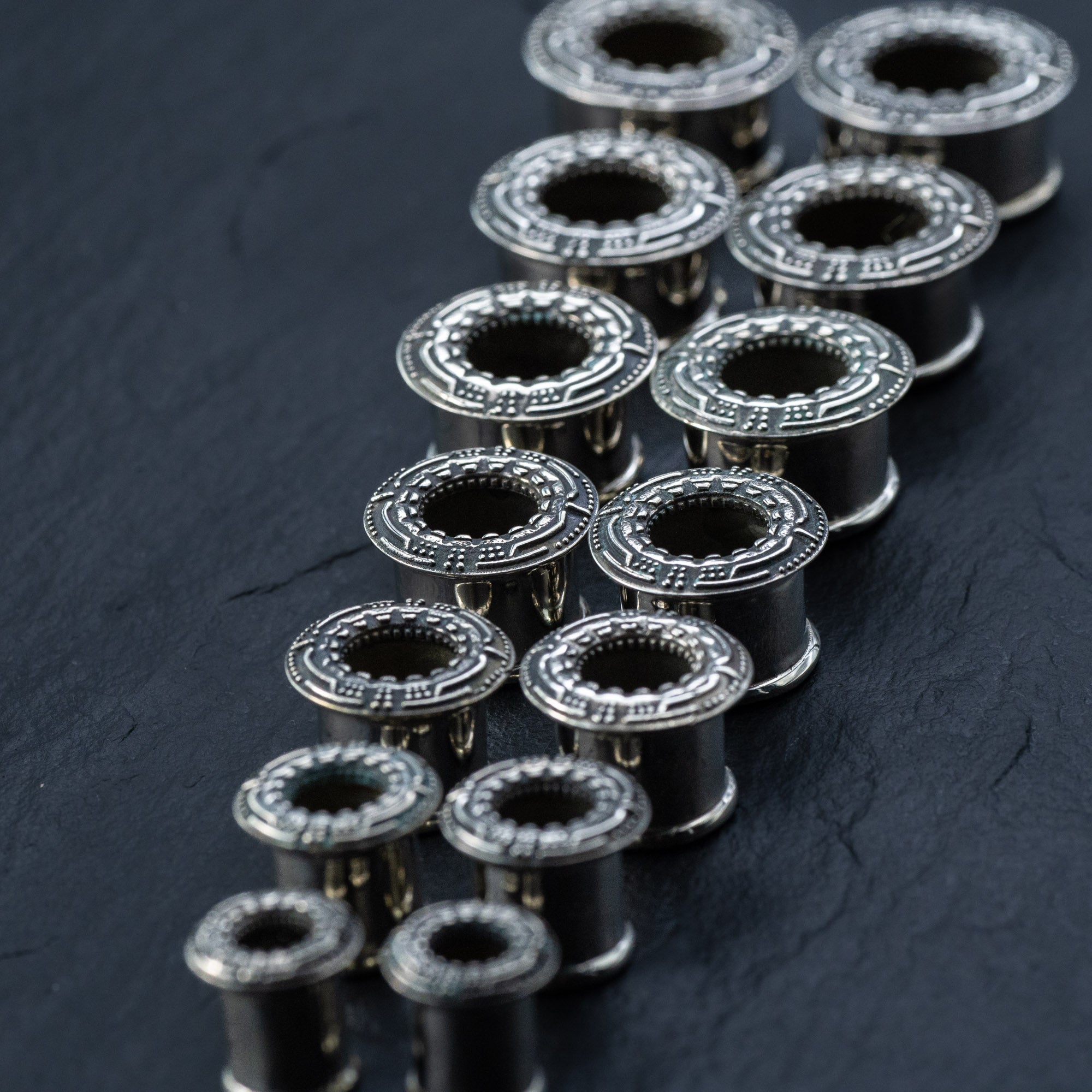 A set of twenty silver brass TERA Futuristic Sci Fi Flesh Tunnels, crafted by Kaya Movement, is arranged on a dark surface. The tunnels, ranging from 8mm to 20mm in size, are lined up diagonally to create a pattern reminiscent of futuristic body jewelry. The lighting highlights their metallic texture and detailed craftsmanship.