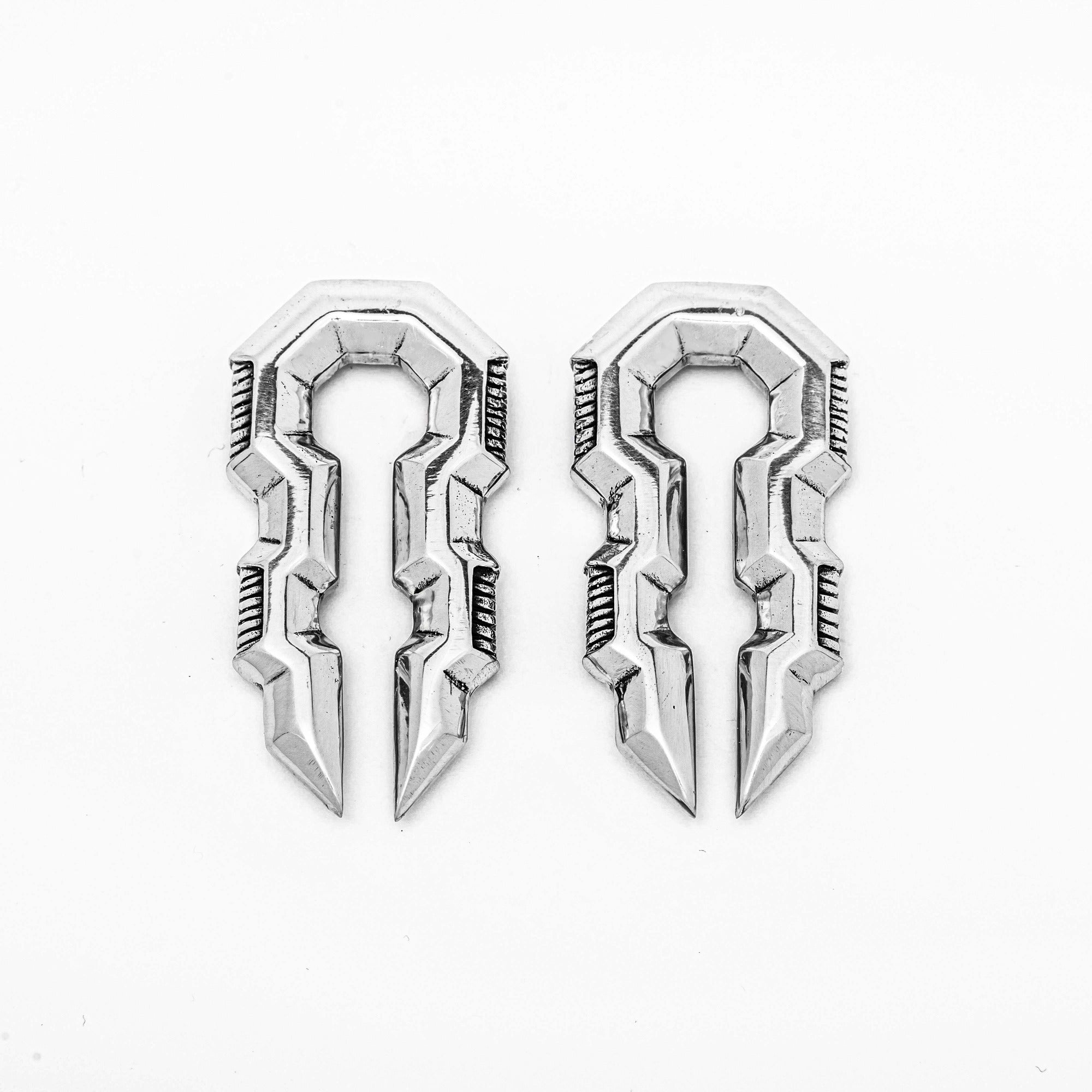A pair of TECHRE Futuristic Hexagon Keyhole Ear Weights in Silver by Kaya Movement, intricately designed with symmetrical, silver-colored, key-like shapes. Each ear weight features sharp, geometric edges and grooves that lend a futuristic and ornamental appearance evocative of post-apocalyptic body jewelry. The 0 gauge ear weights are identical and positioned side by side on a white background.