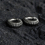 Two dark, ribbed circular TRILOBITE Small Biomechanical Clicker Ear Weights in Silver Brass by Kaya Movement rest on a textured, black surface. These 2 gauge ear weights have a coiled, industrial design reminiscent of biomechanical cyborg earrings and are positioned close to each other, creating a visually striking contrast with the background.