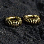 A close-up of the TRILOBITE Small Biomechanical Clicker Ear Weights in Gold Brass by Kaya Movement, resting on a dark, textured surface. The ear weights gleam with a polished finish, and their ribbed design adds a sense of depth and dimension reminiscent of post-apocalyptic body jewelry.