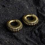 A pair of TRILOBITE Small Biomechanical Clicker Ear Weights in Gold Brass by Kaya Movement lie on a textured dark surface, possibly sand or coarse fabric. The surface's rough texture contrasts with the smooth, shiny finish of the bold, intricately designed 2 gauge tribal earrings.