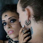 Two individuals with distinctive makeup and body art. The person on the left, who gazes towards the camera, has a crescent moon and intricate lines on their face, enhanced by a nose ring, choker, and biomechanical teardrop hoop earrings. The person on the right sports an ear cuff, neck tattoo, and Kaya Movement's MARVDASHT Medium Cyborg Clicker Ear Weights in Gold (3/18" gauge).