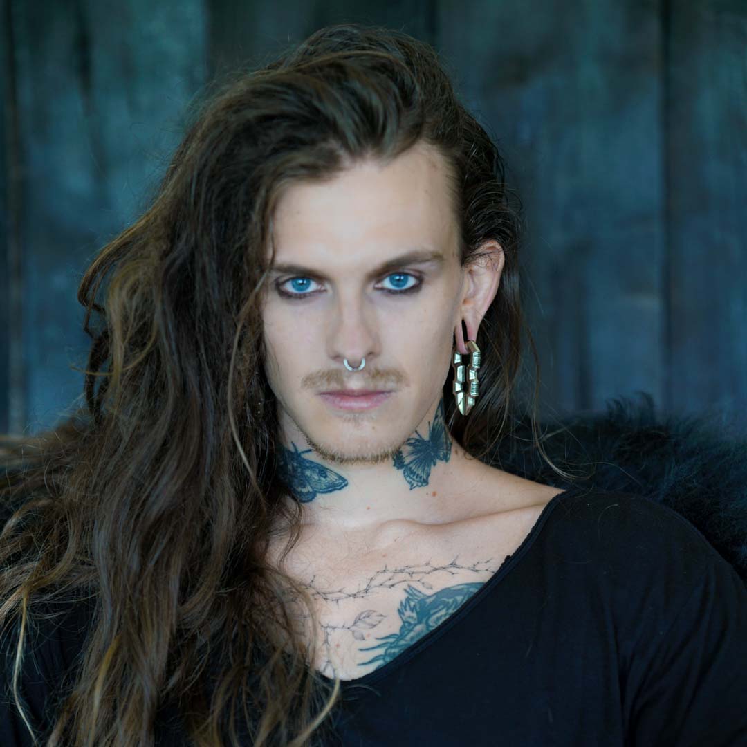 A person with long wavy hair, facial tattoos, a nose ring, and TECHRE Futuristic Hexagon Keyhole Ear Weights in Silver by Kaya Movement is gazing intensely at the camera. They are wearing a black top, and the background is a blurred dark setting, adding a dramatic effect to the portrait.