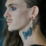 A person with long hair is shown in profile view, flaunting blue eye makeup and a Darma Biomechanical Clicker Hoop Septum Nose Ring in Silver by Kaya Movement. They also sport large ear gauges and a butterfly tattoo on their neck, amidst a blurred background.