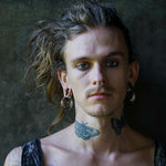 A person with long hair tied back, adorned with PROMETHEUS Medium Biomechanical Hoop Earrings in 925 Silver by Kaya Movement, and showcasing various tattoos – including a butterfly on the neck – as well as facial piercings. The person has a neutral expression and is set against a dark, textured background.