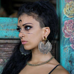 A young woman with long curly hair and a half-shaved head stands sideways, wearing the CIRCUIT Oversized Visionary Disc Hoop Earrings in Gold by Kaya Movement. She also sports a nose ring and necklace, with black geometric and abstract face paint adorning her face, making a striking boho statement. The rustic wooden background decorated with floral accents adds to the overall aesthetic.