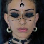A person with dark hair and closed eyes has black face paint designs, including a crescent moon on their forehead and lines across their face. They wear gold brass hoop earrings, an AARO Cyborg Seamless Hoop Septum Nose Ring in Gold from Kaya Movement (18 gauge), and a dark choker necklace.