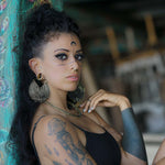 A woman with intricate face and body tattoos, CIRCUIT Oversized Visionary Disc Hoop Earrings in Gold by Kaya Movement adorning her ears, and a nose ring leans against a teal, flower-painted post. She has long, dark curly hair and wears a black top. The background is blurred, suggesting an outdoor or partially open space.