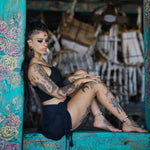 A woman with tattoos sits cross-legged on a rustic wooden ledge, exuding a boho statement. She has long dark hair and is wearing the CIRCUIT Oversized Visionary Disc Hoop Earrings in Gold by Kaya Movement. Dressed in a black crop top and black shorts, she stands out against the backdrop of colorful floral designs and various wooden structures.