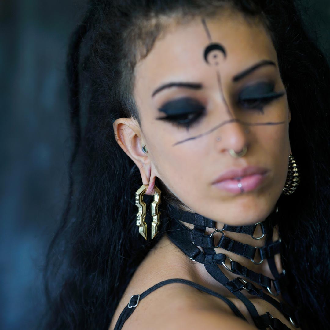 A person with long, dark hair, bold black eye makeup, and distinct facial markings, including a crescent shape on the forehead, is wearing elaborate 0 gauge TECHRE Futuristic Sci Fi Hexagon Keyhole Ear Weights in Gold by Kaya Movement and a harness-like choker. The photo is taken with a blurred background, drawing focus to the person.