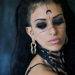 A person with long, dark hair, bold black eye makeup, and distinct facial markings, including a crescent shape on the forehead, is wearing elaborate 0 gauge TECHRE Futuristic Sci Fi Hexagon Keyhole Ear Weights in Gold by Kaya Movement and a harness-like choker. The photo is taken with a blurred background, drawing focus to the person.