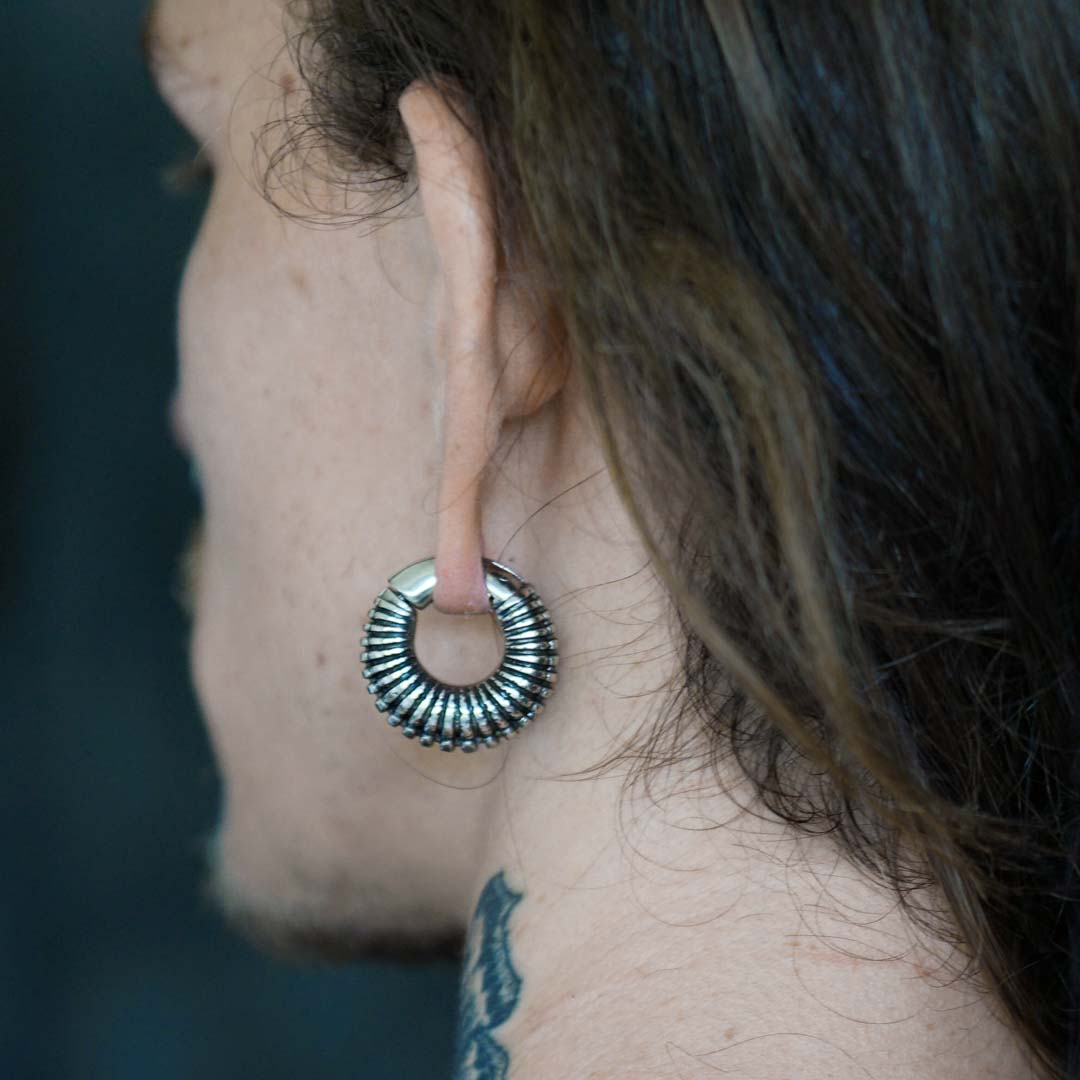 A person with long brown hair is shown from the side, displaying a TRILOBITE Small Biomechanical Clicker Ear Weight in Silver Brass by Kaya Movement. The ear weight, designed to resemble biomechanical cyborg aesthetics, hangs prominently from their 2-gauge earlobe. The person's ear and part of the neck are visible, adorned with a small tattoo above the neckline. The background is blurred and dark.