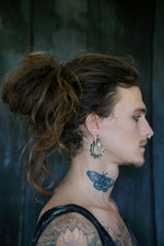 A person with long, curly brown hair in a messy bun, wearing Kaya Movement's GIGER Biomechanical Open Ended Hoop Ear Weights in Silver 925 (0 gauge) and post-apocalyptic body jewelry, has a side profile visible. They have a butterfly tattoo on their neck and a nose ring, displaying a patterned tattoo on their arm against a dark background.