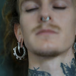 Close-up of a person wearing the GIGER Biomechanical Open Ended Hoop Ear Weights in Silver 925 by Kaya Movement. The large, decorative ear weights feature a pointed claw design reminiscent of post-apocalyptic body jewelry. The individual also has a septum piercing, neck tattoos, short facial hair, and appears to have closed eyes against a blurred background.