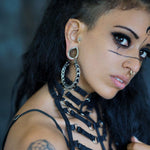 A person with dark hair and piercings, sporting bold makeup and a harness-style top. They are wearing the Quetzalcoatl Oversized Snake Spine Hoop Earrings in Silver White Brass by Kaya Movement. The side of their head is shaved, revealing a tattoo on their shoulder as they look slightly to the side against a blurred background.