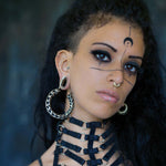 A person with long dark hair and elaborate makeup, featuring a crescent moon on their forehead and lines extending from their eyes. They wear the Quetzalcoatl Oversized Snake Spine Hoop Earrings in Silver White Brass by Kaya Movement and a necklace with multiple straps. The background is blurred and dark.
