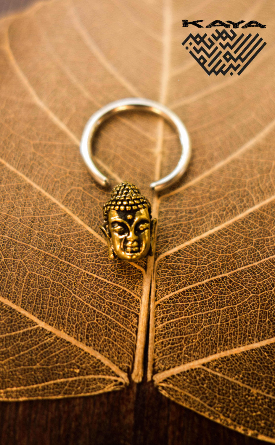 A Buddha silver ring with a gold pendant from Kaya Movement is placed on a large dried leaf with visible veins. The keychain loop is open, showcasing the intricate details of the Buddha's face. The top right corner features the "Kaya Movement" logo in a stylized font.