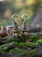 The ANGKOR WAT Heavy Ear Weights in Gold by Kaya Movement, designed with intricate faces at the base and a coiled, elongated structure above, rest on a moss-covered log with a blurred natural background.