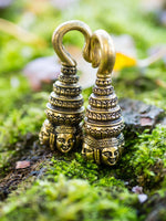 Close-up of ANGKOR WAT Heavy Ear Weights in Gold by Kaya Movement, each featuring detailed Buddha carvings with traditional headdresses, placed on a bed of green moss with blurred background elements. The design showcases intricate patterns and cultural artistry.