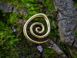 A Brass Spiral heavy ear weight, crafted from nickel-free brass with an antique finish, by Kaya Movement lies on a lush bed of green moss and tree bark, blending into the natural, earthy surroundings. The jewelry's metallic sheen contrasts with the rough, organic texture of the moss and bark.