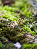 A pair of Tree of Life Stud Earrings in Gold by Kaya Movement lie on a moss-covered log. The intricate tree motifs and 18 gauge craftsmanship stand out against the vibrant green moss, creating a natural, earthy aesthetic. Perfect for ear lobe jewelry enthusiasts, they come in elegant gift packaging.