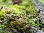 A pair of Tree of Life Stud Earrings in Gold by Kaya Movement, crafted from 18 gauge metal, are placed on a moss-covered log in a forest setting. The background is blurred, highlighting the intricate details of the ear lobe jewelry that resemble small, stylized trees. Ideal for unique gift packaging, these earrings capture nature's elegance.