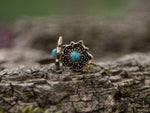 A close-up of the OLLY Mandala Stud Earrings from Kaya Movement, crafted in gold and featuring a choice of turquoise or onyx stones, resting on textured tree bark. The delicate mandala design is complemented by a soft, blurred natural background.
