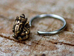 A close-up of a handcrafted GANESHA Silver Ring with Gold Pendant by Kaya Movement, resting on a textured wooden surface. The ring showcases a small, detailed, gold-colored elephant with a raised trunk as the centerpiece. The silver open-ended band contrasts beautifully with the intricate design of the elephant. Available in 18, 16, or 14 gauge.