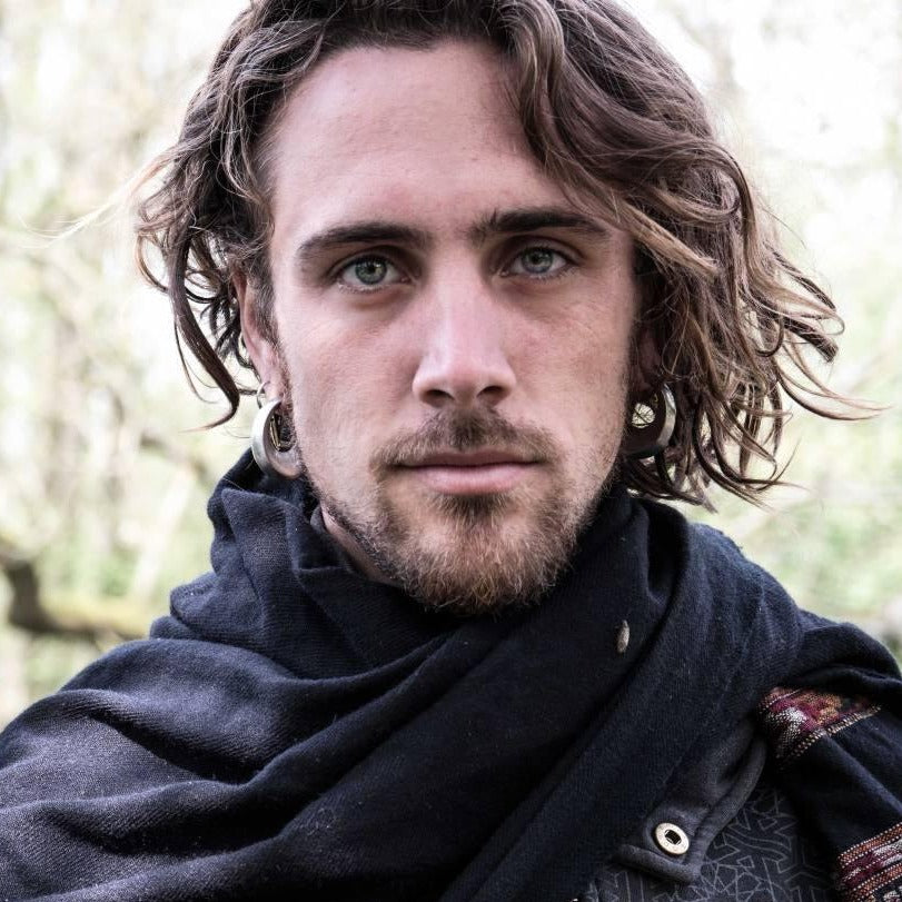 The portrait features a person with wavy, shoulder-length hair and facial hair, adorned with Kaya Movement's HOLO Hoop Dangle Ear Weights in Gold (6 gauge) in their stretched ear lobes. They wear a loose dark scarf or shawl, have light-colored eyes, and gaze directly at the camera with a neutral expression. The blurred background suggests an outdoor setting.