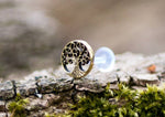 A close-up photograph of the TREE OF LIFE Labret Stud in Gold by Kaya Movement showcases its intricate branches and roots design. The gold brass body jewelry is artfully placed on a piece of bark with green moss, creating a natural and rustic background. The 16 gauge stud earring's metal appears to be bronze-colored.