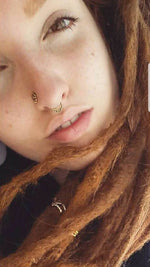 Close-up of a person with light skin and long, reddish-brown dreadlocks. They have light brown eyes and a multi-use piercing adorned with the Kaya Movement LILY Seamless Teardrop Septum Ring in Gold, 18 gauge. Their expression appears neutral or relaxed.