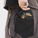 A person is holding a black AMONI Gold Stud Bi-Fold Flip custom leather wallet by Kaya Movement, featuring a decorative labradorite stone, partially tucked into a dark pants pocket. The person has a large silver ring on their finger and a chain attached to their pants.