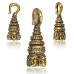 Introducing the ANGKOR WAT Heavy Ear Weights in Gold by Kaya Movement: These 6 gauge brass ornaments are reminiscent of body jewelry, featuring a hook atop a tiered structure adorned with intricate detailing. The base is decorated with multiple expressive faces etched around it, and one angle reveals an elaborate Buddha carving. The image showcases three different views against a white background.