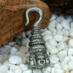 The ANGKOR WAT Heavy Ear Weights in Silver from Kaya Movement is a detailed metal pendant featuring a stacked design with ornate carvings reminiscent of Buddha carvings and multiple faces at the base. Resting on a bed of white pebbles, the pendant showcases a hook-like shape at the top, highlighting its intricate craftsmanship. This piece is perfect for tribal piercing body jewelry enthusiasts and comes in a 6 gauge size.