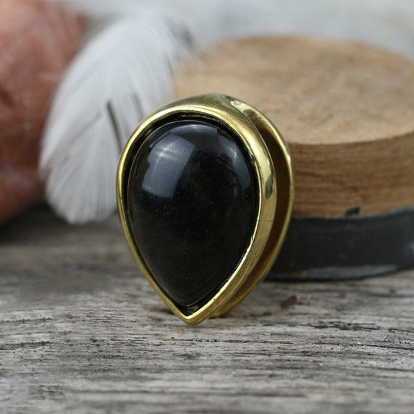 An elegantly designed brooch with a gold frame and golden obsidian stone, much like the Kaya Movement's OBI Minimalist Oval Ear Hangers in Gold & Golden Obsidian, rests on a wooden surface. In the blurred background, a white feather and rustic wooden objects accentuate its sophistication.