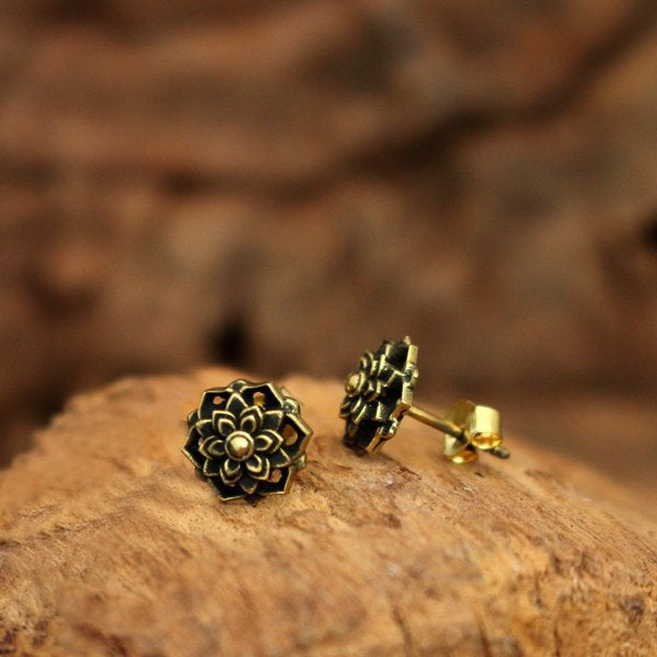 A pair of ILLY Lotus Mandala Stud Earrings in Gold by Kaya Movement, crafted from yellow brass with intricate floral designs, rests on a textured wooden surface. The soft-focused background highlights the detailed craftsmanship of these 18-gauge earrings for your ear lobe.