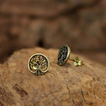 A pair of Kaya Movement's Tree of Life Stud Earrings in Gold (18 gauge), made from yellow brass, are displayed on a textured wooden surface. The ear lobe jewelry features intricate tree designs with roots and branches, capturing a rustic and earthy aesthetic. Perfectly suited for gift packaging.
