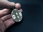 A close-up image of a hand holding the Kaya Movement's ICARO - White Brass Handmade Shipibo Icaros Pendant against a dark background. The pendant features symmetrical, geometric patterns that resemble interlocking cross shapes with oxidized details. The white brass metal has a shiny, reflective surface.