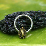 A close-up image of the ornate, handcrafted GANESHA Silver Ring with a Gold Pendant by Kaya Movement, showcasing a detailed elephant design on its front. The 316L steel body jewelry is set against a textured, dark, and reflective background, creating a vivid contrast with the green and black tones.