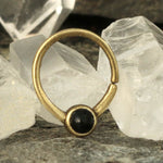 The NAE - Onyx Jewelry by Kaya Movement, an 18g (1mm) ethnic septum ring, is elegantly displayed against a rocky background, nestled between clear quartz crystals. This small gold nose ring features a handmade open-ended design with a captivating onyx gemstone at its center, creating a striking contrast between the gold and black elements.