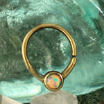 The NAE Seamless Septum Ring in Gold & Opal by Kaya Movement is displayed against a textured glass background, highlighting its natural opal gemstone that showcases a vibrant play of red, blue, and green hues. The slightly blurry background enhances the ring's focus. Ideal for body jewelry or multi-use piercing, the band features an open gap near the gemstone.