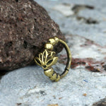 The AVA Lotus Flower Seamless Nose Ring in Gold by Kaya Movement, featuring a lotus flower design and small bead embellishments, rests against a textured rock surface. The intricate details of this 20 gauge body jewelry are highlighted by the natural, rustic background.