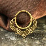 The MANDA Mandala Septum Ring in Gold by Kaya Movement, an 18 gauge piece of gold jewelry, is displayed against a textured brown fabric background on a stone surface. This exquisite body jewelry features ornate geometric and dotted designs with intricate decorative elements at the bottom, exuding a tribal ethnic mandala vibe.