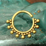 The BRAI Seamless Septum Ring in Gold, crafted by Kaya Movement, boasts a circular gold brass design adorned with small gold beads neatly arranged around the lower half. The backdrop is a textured, teal-colored surface. This ring's tribal ethnic and ornate design offers versatility as a multi-use piercing accessory.
