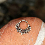 A close-up image of the MANDA Mandala Septum Ring in 925 silver by Kaya Movement, featuring a half-moon shape with detailed dot and line patterns, placed on a reddish-brown, veined stone. The tribal ethnic mandala design stands out against the blurred background, emphasizing the ring's artistry and the stone's texture.