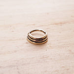 The TRIA Septum Ring in Gold by Kaya Movement, an 18-gauge ring with a minimalist, multi-banded design of three thin stacked bands, rests on a light wooden surface. Its polished finish and sleek, modern appearance evoke the elegance of brass body jewelry.