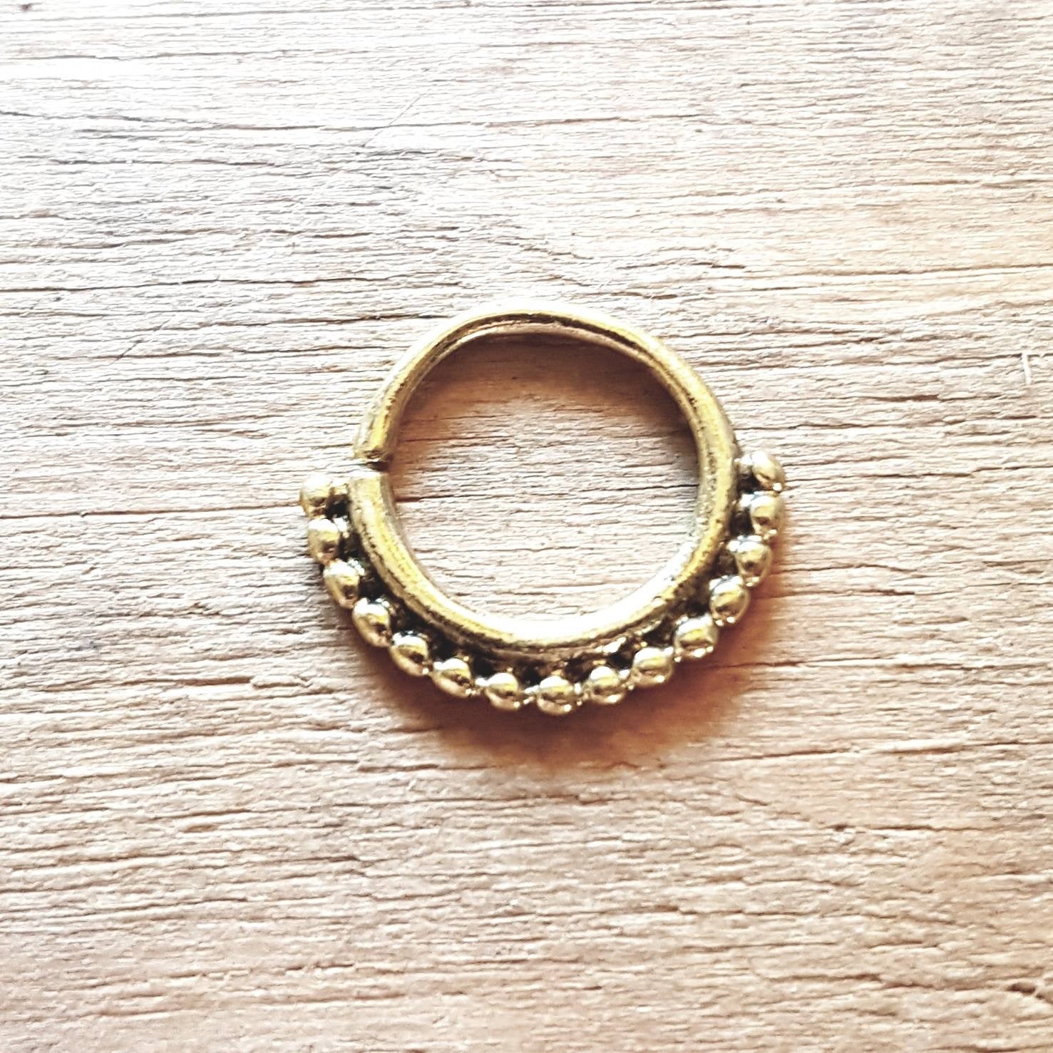 A close-up of the Kaya Movement's IRI Seamless Septum Ring in Gold (16 gauge), adorned with a row of small, evenly spaced beads along the bottom curve, placed on a textured wooden surface, showcasing its tribal ethnic design body jewelry aesthetic.