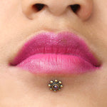 Close-up of a person's lips adorned with bright pink lipstick. Below the lips is a chin piercing featuring Kaya Movement's BEA Flower Labret Stud in Gold & Amethyst, designed with a tribal beaded flower and a natural Amethyst stone at its center. The person's skin tone complements the vivid lip color and the stunning piercing.