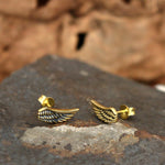 The EL Angel Wings Stud Earrings in Gold by Kaya Movement are showcased on a textured rock against a blurred natural background. These 18-gauge earrings feature intricate wing detailing and a shiny finish, delicately adorning the earlobe. They also come in elegant gift packaging.