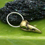 Close-up of a handcrafted jewelry piece from Kaya Movement, known as the CROW SKULL Gothic Silver Ring with Gold Charm. The ring features a gold brass raven skull pendant and is set against a polished, dark green, textured stone background. The band is made of smooth 316L surgical steel, while the detailed bird skull charm rests on a green surface. Available in 18, 16, or 14 gauge sizes.